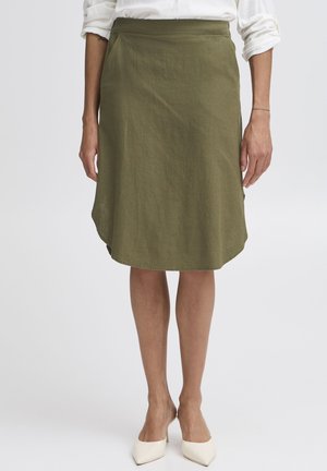 Kobieta ubrana w oliwkową spódnicę do kolan, białą bluzkę z podwiniętymi rękawami i białe szpilki, stojąca na jednolitym tle.