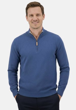 Man wearing a blue long-sleeve quarter-zip sweater with hands in pockets, smiling against a plain light background.