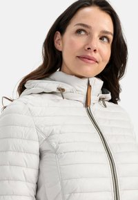 Letvægts, hvid pufferjakke med vandret syninger, lynlås og hætte. Har snortræk og brune detaljer på hardware.
