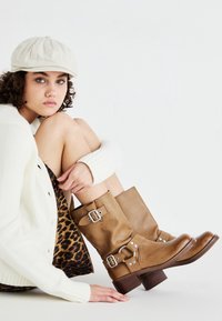 Ung kvinna med lockigt hår klädd i en beige keps, vit tröja, leopardmönstrad kjol och bruna läderstövlar med spänne, sittande mot en vit bakgrund.