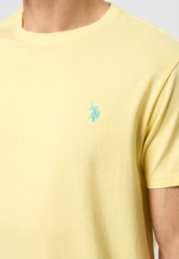 Tricou din bumbac galben cu guler rotund, având un mic logo brodat de culoare albastru deschis pe partea stângă a pieptului și mâneci scurte.