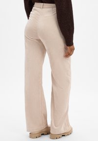 Pantalons en velours côtelé beige avec une texture côtelée, jambes larges et poches arrière, associés à des chaussures à plateforme beiges. Vue arrière montrée.