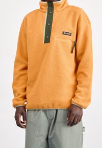 Orange fleecetröja med hög krage, med gröna tryckknappar, en framficka och kontrasterande krämfärgade detaljer längs kragen och ärmsluten.