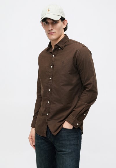 Hombre con camisa marrón de botones, jeans oscuros y gorra de béisbol beige, de pie con una mano en el bolsillo contra un fondo blanco liso.
