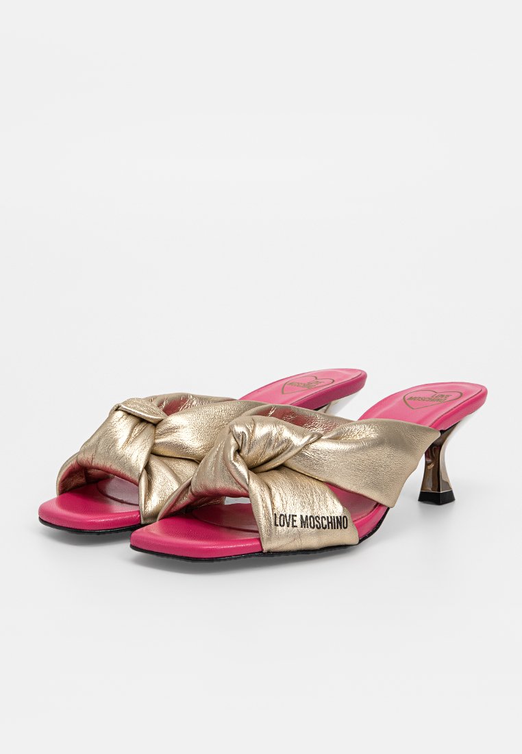 Goldene metallic High-Heelsandalen haben ein knotendes Design an der Vorderseite, pinke Innensohlen und einen kleinen, durchsichtigen Blockabsatz. Die Markenbezeichnung "LOVE MOSCHINO" ist sichtbar.