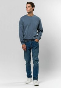 Sivý sveter s okrúhlym výstrihom, rebrovanými manžetami a malým logom na hrudi, skombinovaný s slim-fit modrými džínsami a bielymi teniskami.