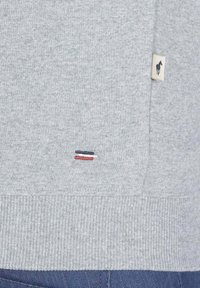 Sweatshirt gris avec un ourlet côtelé, présentant un petit détail brodé de trois lignes horizontales en marine, rouge et blanc, ainsi qu'un patch logo.