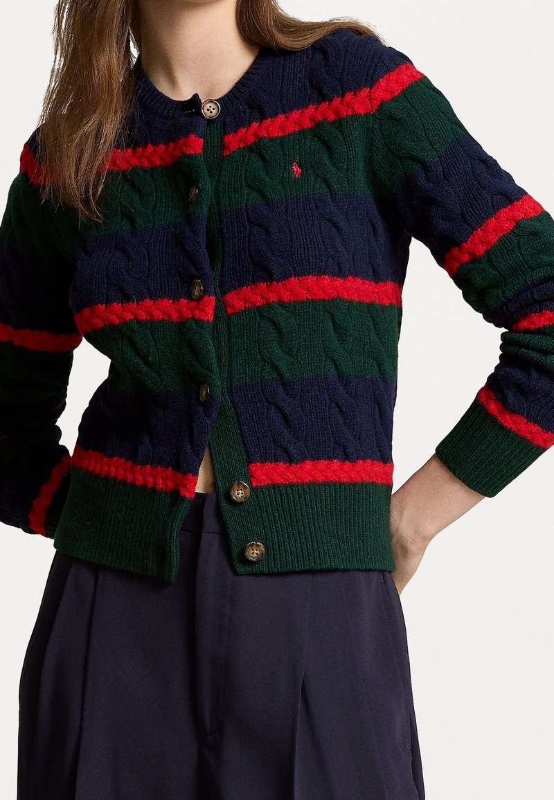 Femme portant un cardigan en maille torsadée à rayures bleu marine, vert et rouge avec des boutons, et un pantalon taille haute sombre, bras plié à la taille.