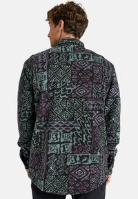 Camisa de manga larga en tejido oscuro con patrones geométricos en azul verdoso y púrpura. Presenta un cuello tipo camisa y puños abotonados. Textura suave.