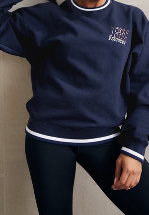 Person iført navy blå PE Nation sweatshirt med hvid kant og sorte leggings, stående mod en ensfarvet beige væg.