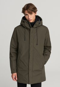 Giacca imbottita verde oliva con cappuccio, caratterizzata da chiusura a zip, cordini e tasche laterali. Tessuto liscio, design di lunghezza media.