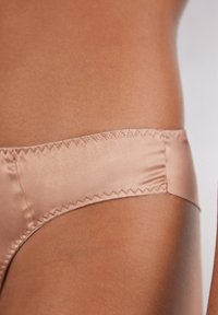 Satinowy string w kolorze nude o gładkiej, miękkiej fakturze. Posiada zygzakowe przeszycie wzdłuż pasa i minimalistyczny design.