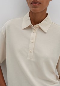 Polo beige en tissu texturé, avec un col classique et une patte de boutonnage à quatre boutons. Manches courtes avec un détail de couture subtil.