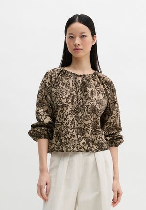 Frau mit glattem schwarzen Haar trägt eine braune Bluse mit floralem Muster, gerafften Ärmeln und lockeren weißen Hosen vor einem schlichten Hintergrund.