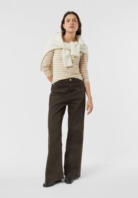 Kobieta stoi, ubrana w beżowo-białą bluzkę z długim rękawem w paski, kremowy sweter zarzucony na ramiona, ciemne spodnie o szerokich nogawkach i czarne buty.