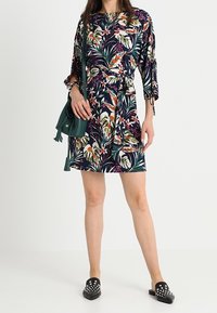 Vestido de estampado floral con un fondo azul marino oscuro, mangas de corte suelto, cinturilla con lazo y un corte a la altura de la rodilla. Combinado con un bolso verde y sandalias negras.