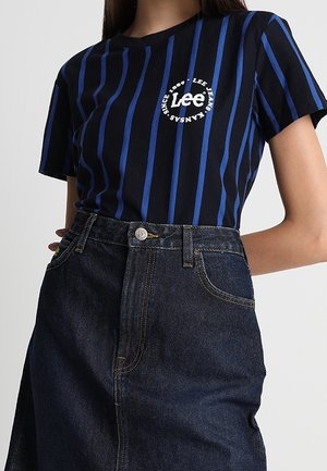 Kobieta ubrana w czarno-niebieską, paskowaną koszulkę Lee Jeans, wpuszczoną w wysokowarstwową, ciemną spódnicę dżinsową z guzikiem i kieszeniami.