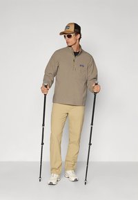 Smėlio spalvos megztinis su ketvirčio užtrauktuku, šviesios smėlio spalvos kelnės, dėvi smėlio spalvos kepurę ir saulės akinius, laiko žygių lazdas, turi elegantišką sportinį dizainą.