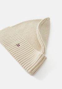 Căciulă tricotată bej, cu o textură ribbed și cu manșetă pliată. Prezintă un mic emblemă albastră, albă și roșie pe lateral.