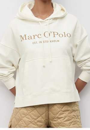 Biała bluza z kapturem z napisem "Marc O'Polo EST. IN STOCKHOLM", zestawiona z beżowymi pikowanymi szortami, ręce opierające się na biodrach.