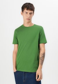 Green cotton t-shirt with a crew neck and short sleeves. The fabric appears soft, with a smooth texture and a relaxed fit. A tattoo is visible on the arm.