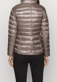 Kvinna som bär en figurnära, blänkande taupe-färgad vadderad jacka med långa ärmar och svarta byxor, sedd från baksidan mot en neutral bakgrund.