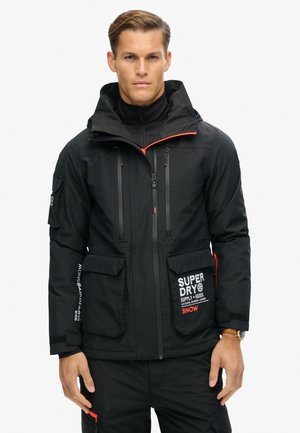 Sort, isoleret jakke med hætte, med røde lynlåsdetaljer, flere lommer og "SUPERDRY" branding. Blødt, struktureret stof.