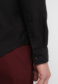 Svart texturerad långärmad skjorta med knappade manchetter, kombinerad med vinröda byxor. Skjortan har en rak fåll och synliga sömdetaljer.