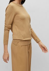 Sweter w kolorze kamelowym, dziany, z długimi rękawami i okrągłym dekoltem. Posiada boczne rozcięcia i prążkowane mankiety, w zestawie z dopasowanymi spodniami.