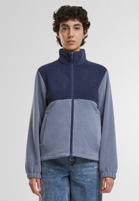 Fleecejakke med høj krave, lynlås foran, farveblokeret design i marineblå og lys grå, med elastiske manchetter og to sidelommer.