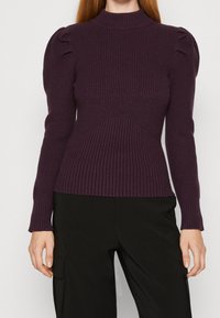 Fialový žebrovaný svetr s vysokým límcem, s nafouklými rukávy a přiléhavým střihem. Texturovaná látka se šikmým pleteným vzorem na přední straně.