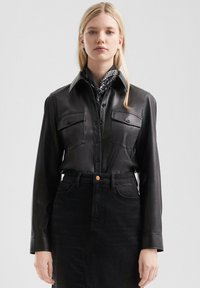 Zwarte leren shirt met lange mouwen, gestructureerde kraag, twee borstzakken en knoopsluiting; gecombineerd met een zwarte denim rok.