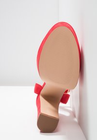 Escarpin rouge en suede avec un bout pointu, une semelle en caoutchouc beige dotée de rainures horizontales et un détail de sangle à la cheville.