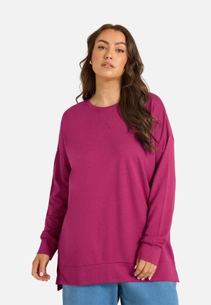 Kvinde med langt mørkt hår iført en løs magenta sweatshirt og lyseblå bukser mod en ensfarvet baggrund.