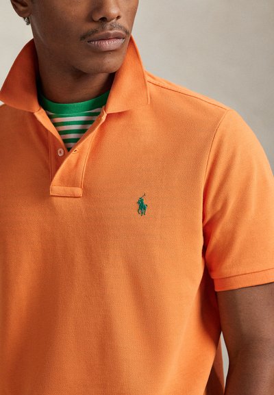 Πορτοκαλί πόλο με υφασμάτινη υφή, διπλωμένο γιακά και πράσινο κεντημένο λογότυπο. Φορεμένο πάνω από πουκάμισο με πράσινες και λευκές ρίγες.
