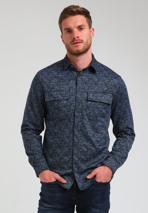 Camicia blu navy con bottoni, con un motivo strutturato, maniche lunghe e due tasche sul petto. Presenta bottoni abbinati e un orlo arrotondato.