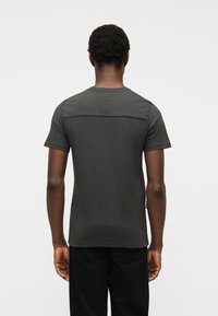 Άνδρας με γυρισμένη την πλάτη, φοράει γκρι σκούρο κοντομάνικο πουκάμισο με οριζόντια ραφή στο πάνω μέρος της πλάτης και μαύρο παντελόνι.