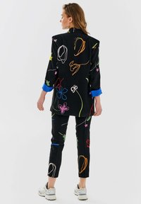 Czarny garnitur z kolorowym abstrakcyjnym haftem; składa się z długiego żakietu z niebieskimi mankietami i wąskich spodni, wykonany z gładkiego materiału.