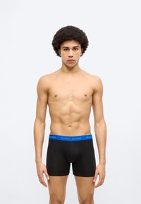 Μαύρα μποξεράκια με μπλε μέση, που φέρουν το λογότυπο "TOMMY HILFIGER". Το υλικό φαίνεται λείο και ελαστικό. Το μοντέλο στέκεται με ουδέτερο έκφραση.