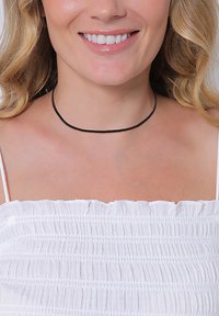 Black choker necklace, thin and smooth, worn around the neck. Model has wavy hair and is dressed in a white smocked top. No additional details.