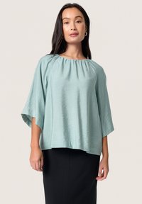 Blusa turchese chiaro con scollo rotondo, maniche drappeggiate e una finitura leggermente testurizzata, abbinata a una gonna nera.
