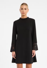 Schwarzes Kleid mit langen Ärmeln, das Spitzenakzente an den Manschetten aufweist. Hoher Halsausschnitt, fließende A-Linie und glatte Stofftextur.