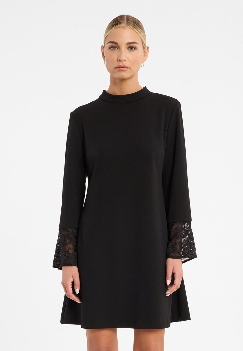 Schwarzes Kleid mit langen Ärmeln, das Spitzenakzente an den Manschetten aufweist. Hoher Halsausschnitt, fließende A-Linie und glatte Stofftextur.