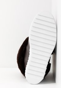 Zapatilla marrón con suela de goma blanca. Cuenta con una parte superior de felpa suave con un acabado liso, diseñada para brindar comodidad y calidez. Suela con agarre texturizado.