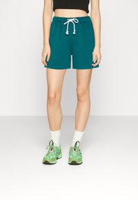 Tyrkysové bavlněné šortky s elastickým pasem a bílým stahovacím páskem, volného střihu, v kombinaci se zelenými teniskami a bílými ponožkami ke kotníkům.