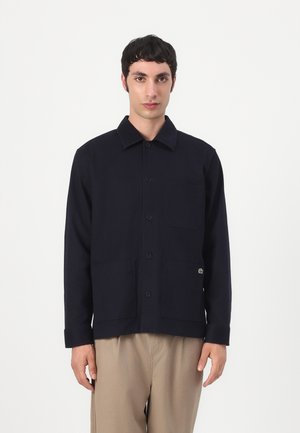 Uomo con capelli corti e scuri che indossa una giacca nera con bottoni e pantaloni beige, in piedi davanti a uno sfondo bianco uniforme.