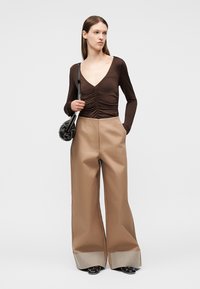 Woman wearing a brown ruched V-neck top, wide-legged beige pants with checkered cuffs, black shoes, and holding a black handbag.