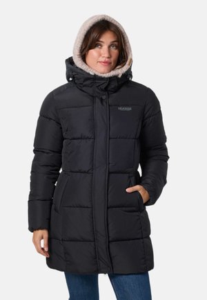 Une femme présente un long manteau d’hiver noir matelassé, doté d’une capuche doublée de polaire, devant un fond uni et clair.