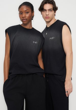 Dve manekenki nosita črne brezrokavnice z majhnim belim logotipom, stojita blizu skupaj na enostavni svetli podlagi.