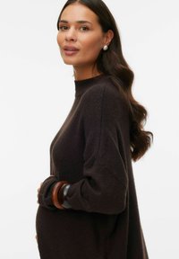 Mørkebrunt strikket sweater med høj hals og løs pasform, der har store træarmbånd på armen, glat tekstur og subtile fibre.